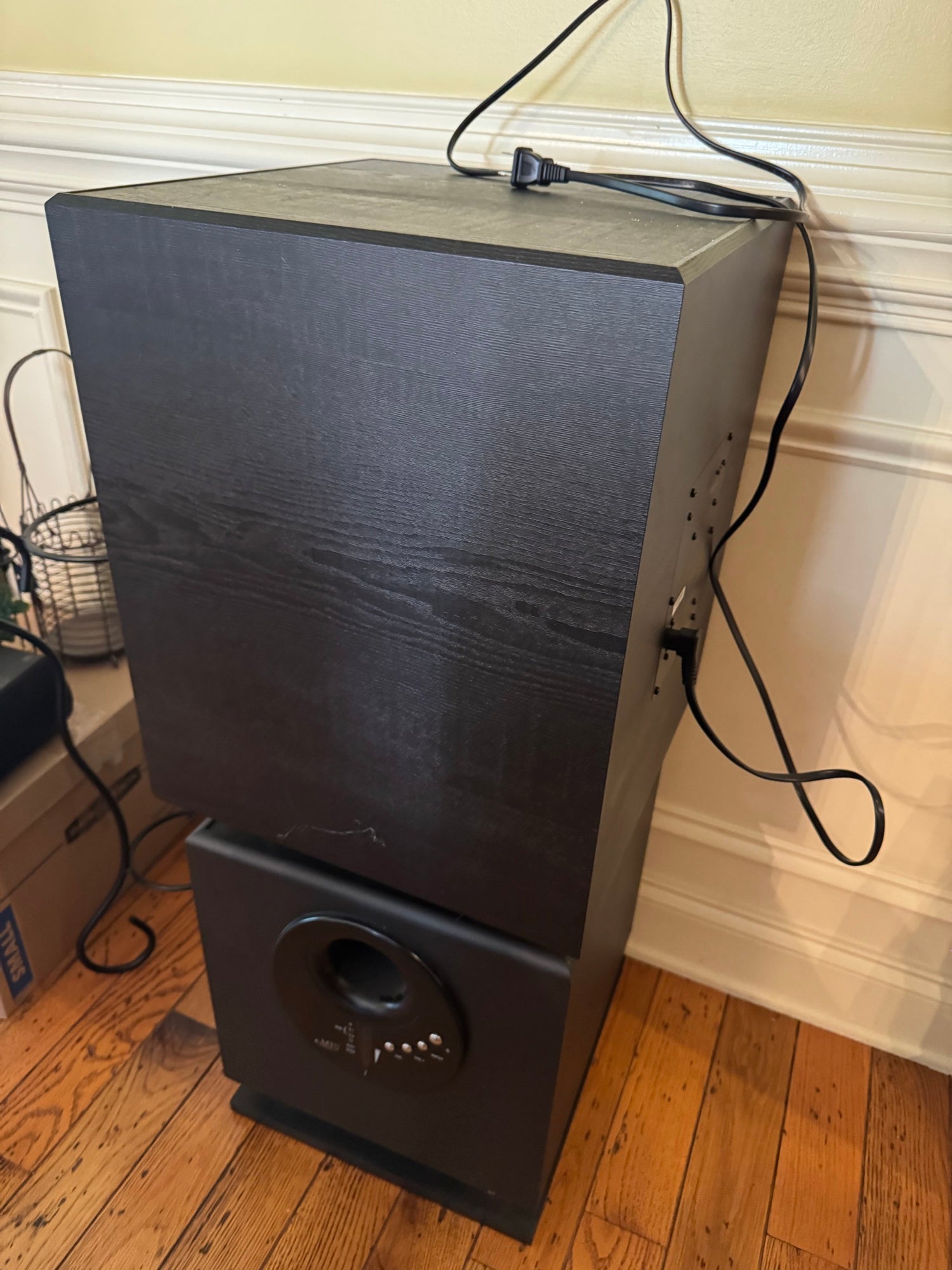 Black subwoofer with textured finish, standing upright on wooden floor against cream wall