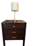 Espresso finish three-drawer nightstand with brushed nickel bar pulls, positioned beside a bed with a lamp on top.