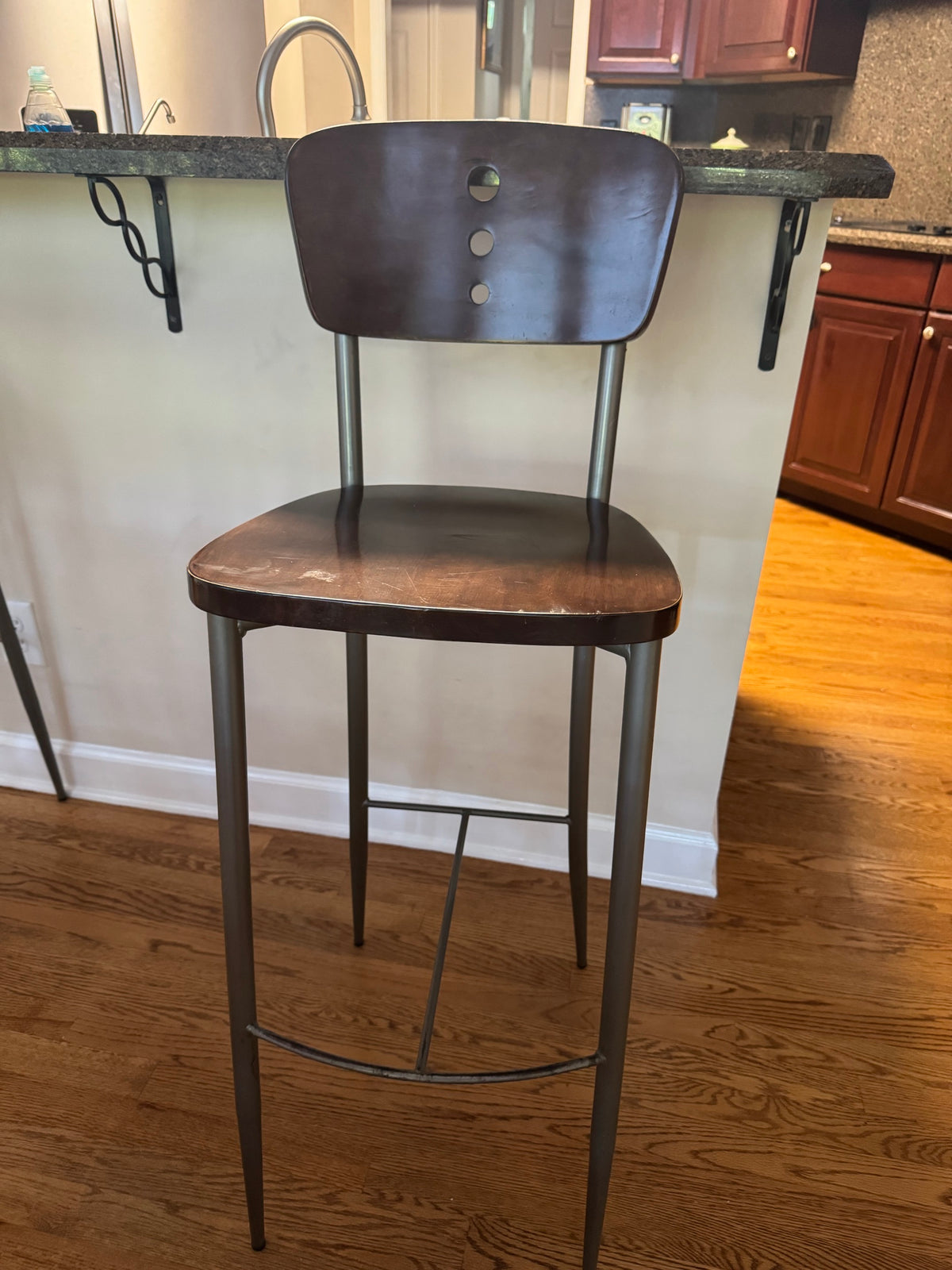 Single bar stool with three-hole cutout wood back and scuffed wood seat, front view against kitchen counter