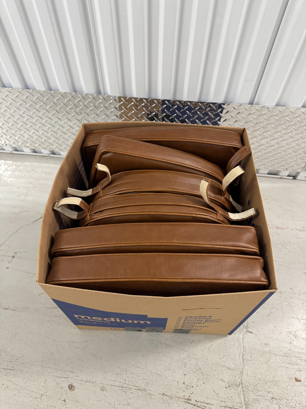 Open cardboard box containing stack of six brown padded dining chair cushions with fabric ties visible