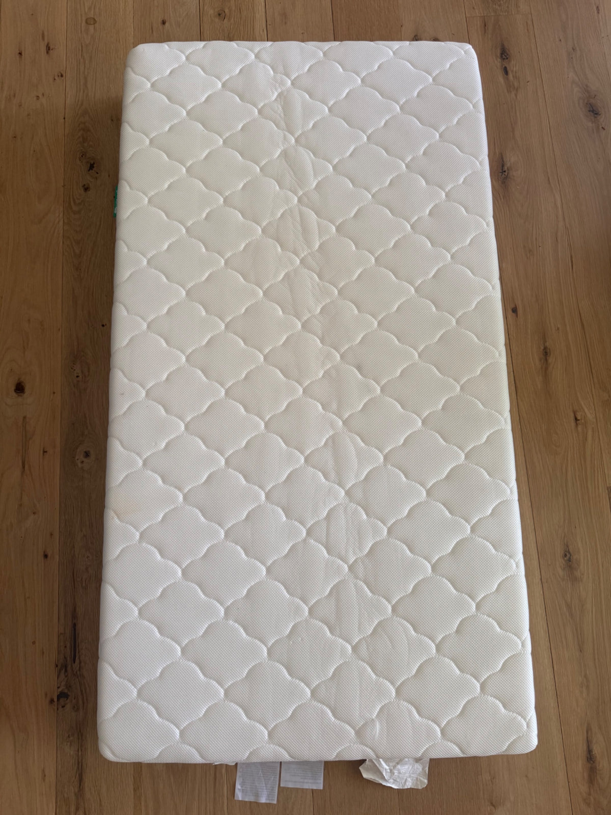 White quilted crib mattress with diamond pattern, photographed flat on wooden floor from directly above