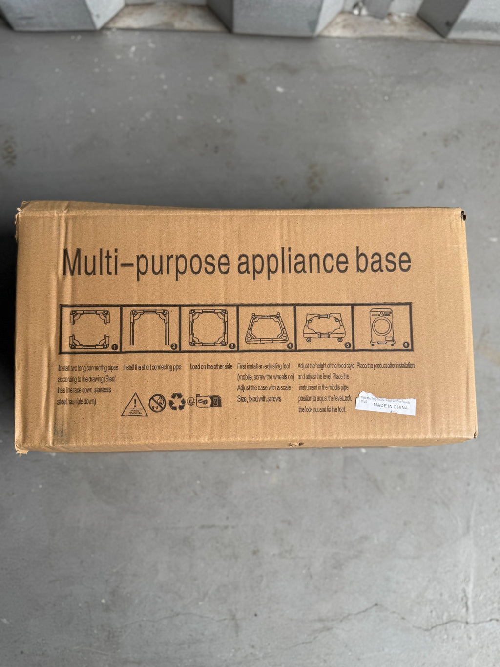 Unwrapped cardboard box on concrete floor showing front label with installation diagrams, warning symbols, and 'Made in China' sticker