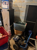 Gas-powered pressure washer with blue and black frame and brass pump, stored among bins and household items in a garage