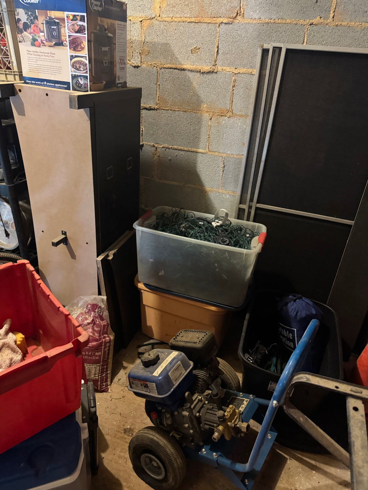Gas-powered pressure washer with blue and black frame and brass pump, stored among bins and household items in a garage