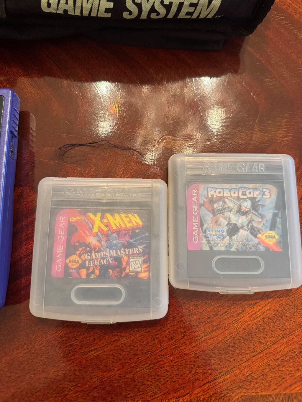 Two Sega Game Gear cartridges in cases, X-Men GamesMaster's Legacy and RoboCop 3, next to the carrying pouch