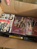 Cardboard box open displaying collection of DVD and Blu-ray cases with various film titles visible, hand holding box flap