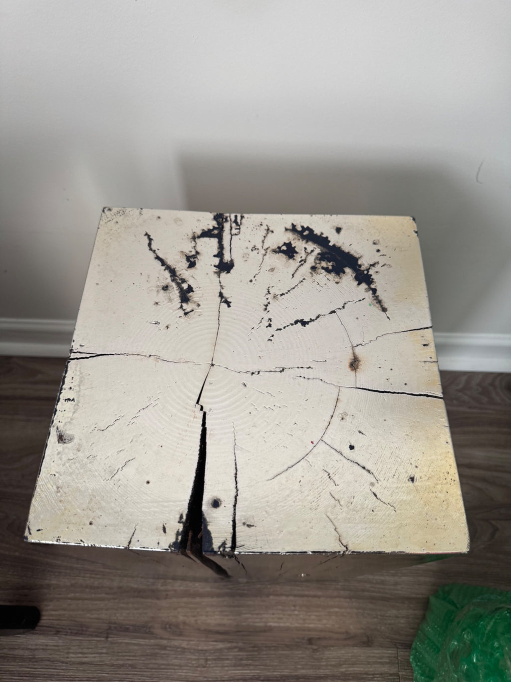 Top-down view of square wooden table top, painted white with black paint splatter and burn marks, showing four visible wood sections meeting at center