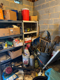 Garage storage corner with metal shelving holding boxes, chemicals, a cooler jug, and yard tools including shovels, a rake, and a gas-powered trimmer