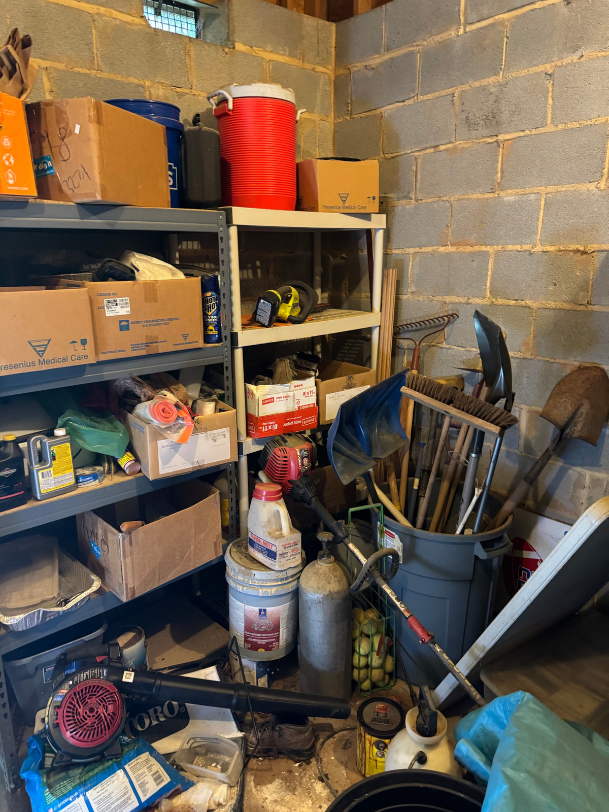 Garage storage corner with metal shelving holding boxes, chemicals, a cooler jug, and yard tools including shovels, a rake, and a gas-powered trimmer